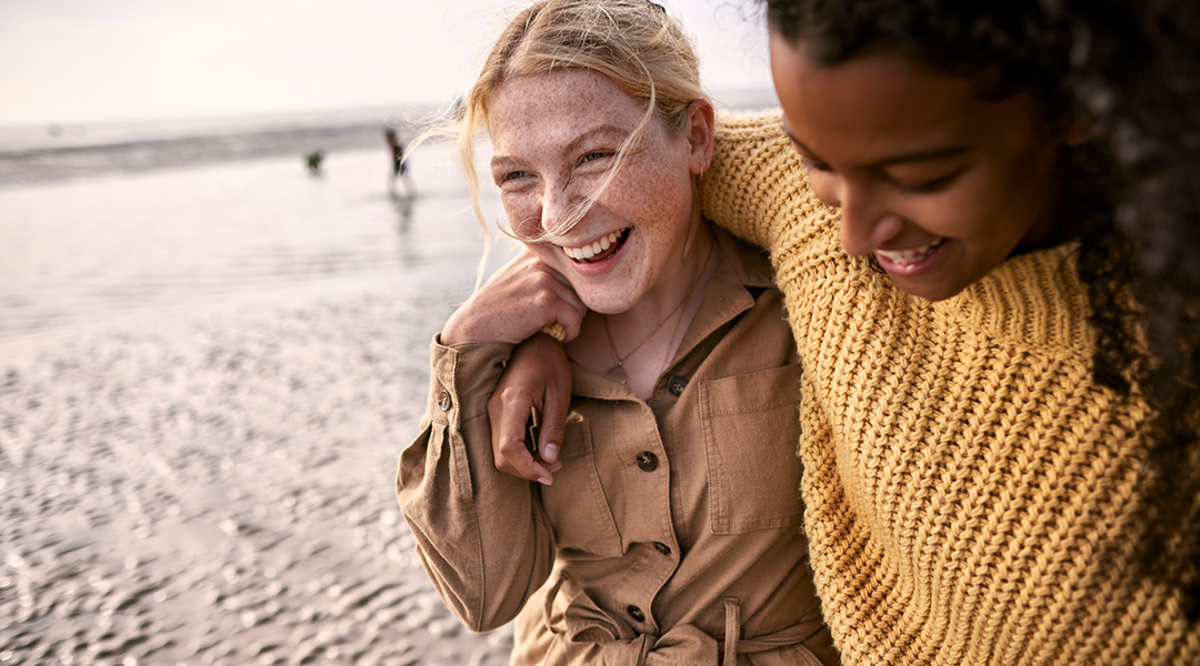 Two girls. One with their arm around the others neck smiling and having a good time on the beach